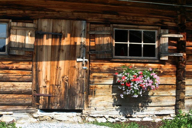 Drevená chata so starým oknom, veľkými dverami a muškátmi na stene.jpg