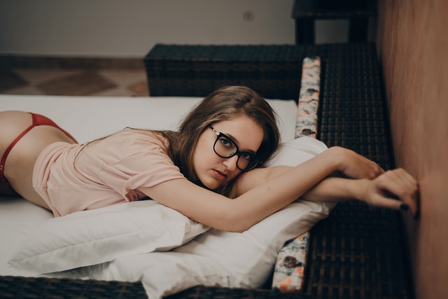 Žena v červených nohavičkách tričku a v okuliaroch leží na bruchu na posteli